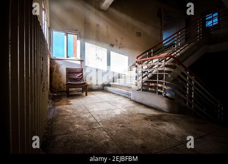 Old hospital building abandoned one single empty chair with eerie creepy sun light rays shining in on dark old haunted decaying staircase landing Stock Photo