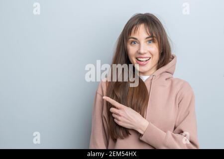 Portrait of a beautiful young brunette woman in a hoodie pointing finger at blank copy space presenting your product. Stock Photo