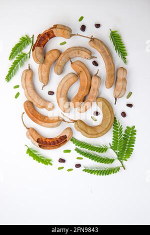 Tamarind  bean like pods filled with seeds surrounded by a fibrous pulp arranged in a white textured background with its fresh green leaf. Stock Photo