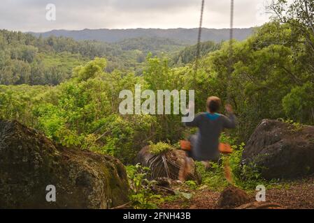 Woman sitting on rope swing hanging from tree, perched on hilltop of Gunstock Ranch, at sunset, Laie, Oahu, Hawaii, USA Stock Photo