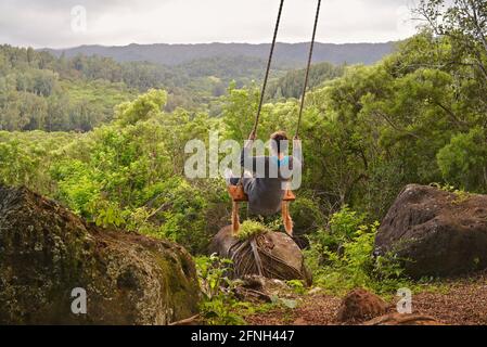 Woman sitting on rope swing hanging from tree, perched on hilltop of Gunstock Ranch, at sunset, Laie, Oahu, Hawaii, USA Stock Photo