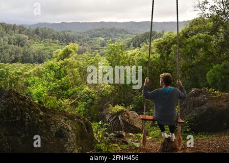 Woman sitting on rope swing hanging from tree, perched on hilltop of Gunstock Ranch, at sunset, Laie, Oahu, Hawaii, USA Stock Photo