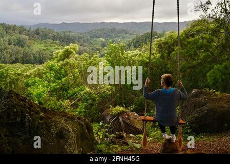 Woman sitting on rope swing hanging from tree, perched on hilltop of Gunstock Ranch, at sunset, Laie, Oahu, Hawaii, USA Stock Photo