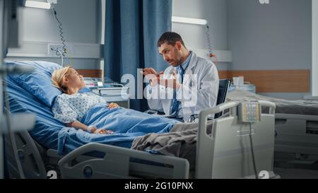 Hospital Ward: Friendly Doctor Talks to Smiling Young Woman Resting in Bed, Fully Recovering after Sickness. Physician Uses Tablet Computer, Does Stock Photo