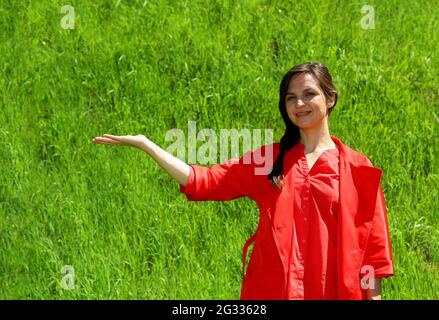 A smiling girl in red clothes on a background of green grass, a hand gesture in the side. A young, confident woman in the background of the green lawn raised her hand to the side. Space for advertisin Stock Photo