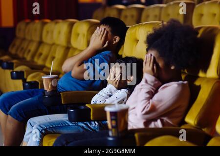 Young boy and girls sitting in movie theater with cold drinks closing their eyes with hands while watching a scary scene of a horror film Stock Photo