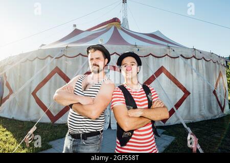 Muscular build artist standing with arms crossed by clown in front of circus tent Stock Photo