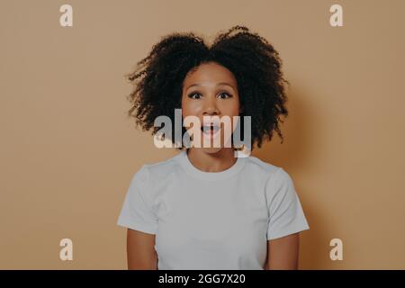 Amazed stunned dark skinned female looking at camera with opened mouth isolated over beige wall Stock Photo