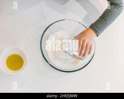 Top view of crop anonymous child pouring water from glass cup on flour in bowl placed on white table near bowl with oil while cooking pastry Stock Photo