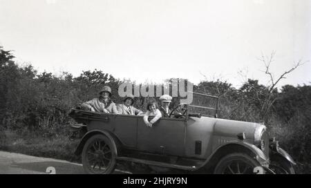 circa 1920s, historical, on a country lane, a family... mother, sister and young daughter and boy, in a cloth cap, sitting in an open-top motorcar of the era, England, UK. Both ladies are wearing hats. A starting handle can be seen sticking out from underneath on the right panel of the car on the driver's side and the horn is next to the steering wheel. Stock Photo