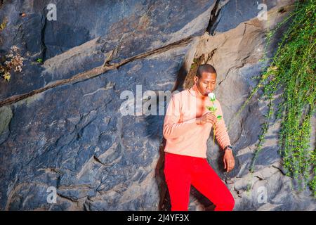 Man Missing You. Dressing in light orange sweater with high collar, red pants, wearing wristwatch, a young black guy is standing against rocks with lo Stock Photo