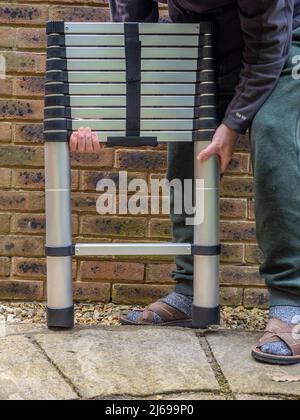 Man holding Aluminium tubular section telescopic access ladder collapsed and strapped together with hook and eye strap Stock Photo