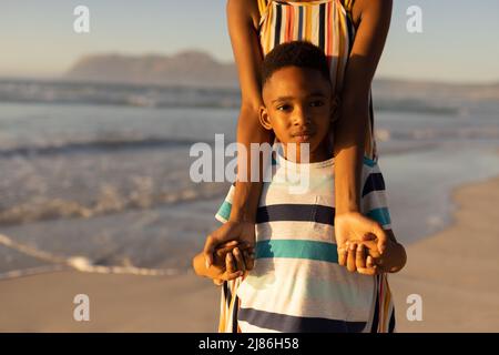 Portrait of african american cute boy holding young mother's hands while standing against sea Stock Photo