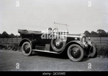 1938, historical, a young school boy, with school cap on, sitting for a photo in his father's touring car which is parked on a coutryside road, England, UK. Possibly made in the 1920s, the open-top car has a simple squeeze horn on the outside beside the driver and a can of motor spirit mounted on the surround underneath the driver's door.  Unusually, the wheels are solid, whereas most wheels had spokes in this era. The spare wheel has the letter S in its hub. Stock Photo