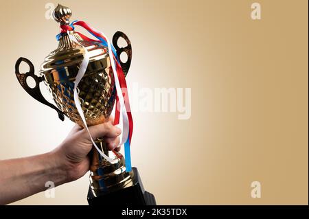 Awards for victories, achievements in sports - a gold cup and medals on multi-colored ribbons in a person's hand on a beige background. Award ceremony Stock Photo