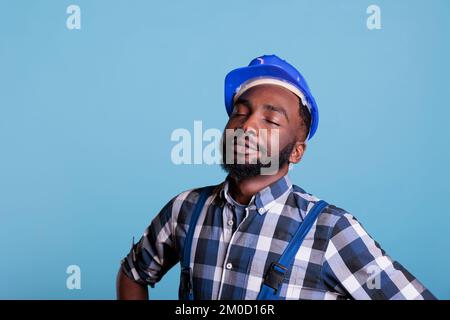 Construction worker with a dream to start work very early in the morning. African american contractor wearing hard hat and work uniform with eyes closed. against blue background, studio shot. Stock Photo