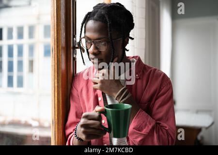 Thoughtful black man looks out window touches chin holding cup thinks about important work matters. Stock Photo