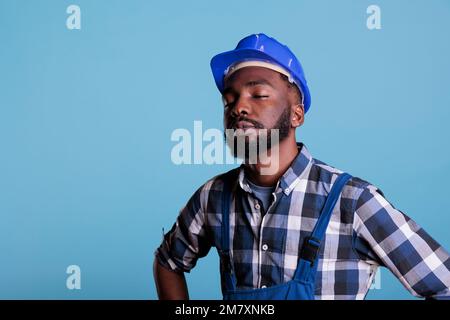 Tired exhausted construction worker in construction helmet and coveralls. African american employee with closed eyes feeling fatigued after hard work isolated on blue background. Stock Photo