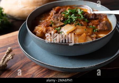 Homemade beef stew with cabbage and vegetables. Served in a grey bowl with spoon on dark wooden and rustic table background. Closeup and front view Stock Photo