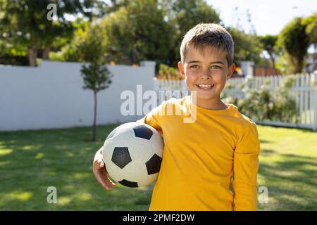 Portrait of cute caucasian boy smiling and holding soccer ball while standing on grassy land in yard Stock Photo