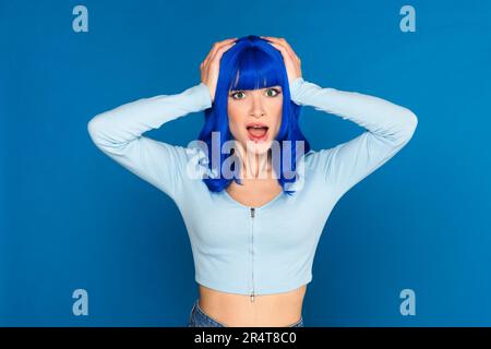 Shocked young female model with dyed bright blue hair in casual clothes grabbing head and looking at camera with opened mouth in studio Stock Photo