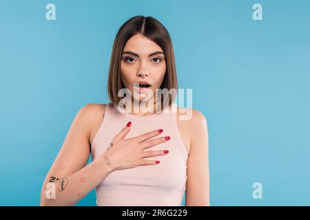 shocked woman with hand on chest, stylish model with tattoos and nose piercing looking at camera on blue background, emotional, opened mouth, generati Stock Photo