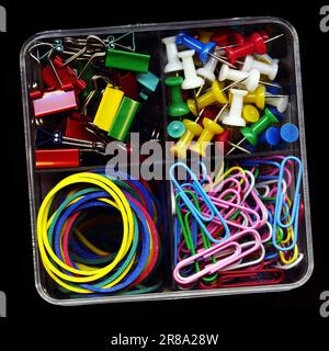 Set of colorful stationery, push-pins, clips, rubber bands, clamps isolated on black. Various office supplies in a plastic box close-up Stock Photo