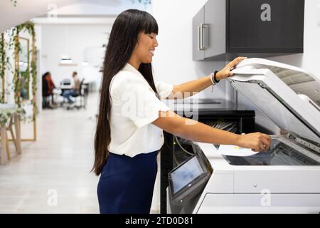 Side view of smiling Asian young businesswoman in casuals with long hair using modern copier while working in creative office Stock Photo