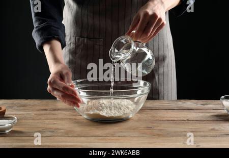 Making bread. Woman pouring water into bowl with flour at wooden table, closeup Stock Photo