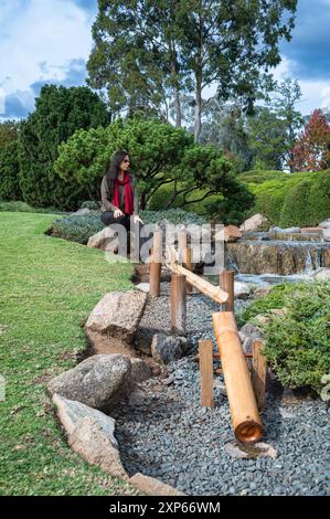 Low angled view of a bamboo water feature leading up to a female tourist, in reflection, seated on a rock near a small waterfall in a Japanese Garden. Stock Photo