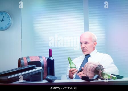 White male in a corporate setting examines a beer bottle, with multiple alcoholic beverages spread on his desk Stock Photo