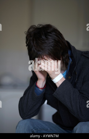 young dark haired teenage man sitting with his head in his hands hiding his face staring at the floor in the kitchen Stock Photo