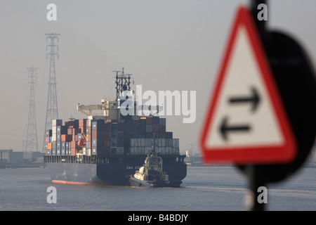 Past a two-way sign, a giant cargo container ship is guided by tug, easing upstream on the River Thames towards Tilbury Docks Stock Photo