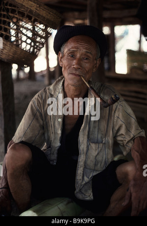 An old man from the Akha tribe of Northern Thailand smokes his pipe sitting under his house Stock Photo