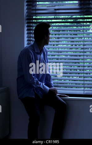 Young man sitting in a dark room looking out through a window blind. Stock Photo