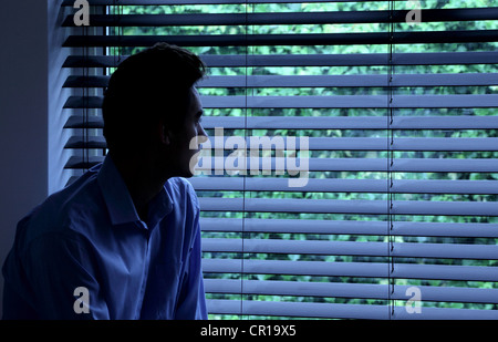 Young man sitting in a dark room looking out through a window blind. Stock Photo