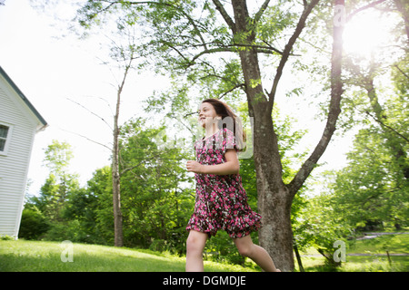 A young girl in a patterned summer dress, running across the grass under the shade of trees in a farmhouse garden. Stock Photo