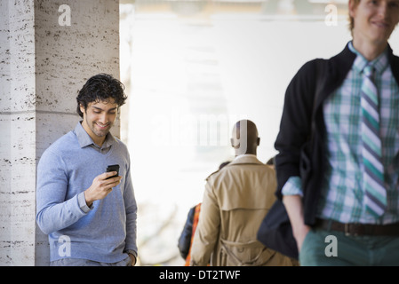 Three people on the sidewalk a man using his mobile phone a man in a raincoat and a young man walking along the street Stock Photo