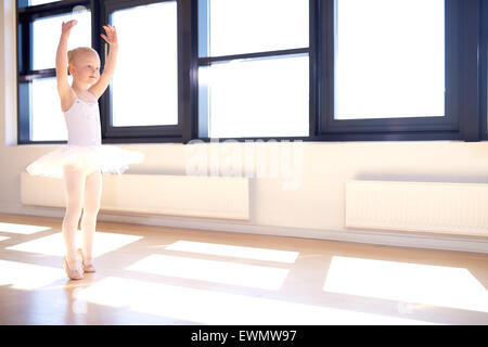 Little girl training to be a ballerina standing in a graceful arms raised position in her white tutu and pink satin ballet shoes Stock Photo