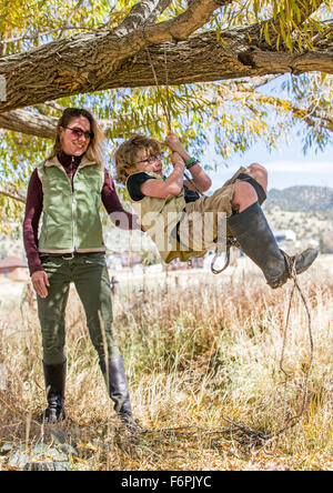 Attractive mother helps young son on rope swing hung from ranch tree Stock Photo