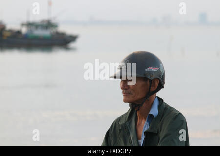 Vietnamese man with motorbike helmet in front of fishing boat and South China Sea at Danang beach, Vietnam Stock Photo