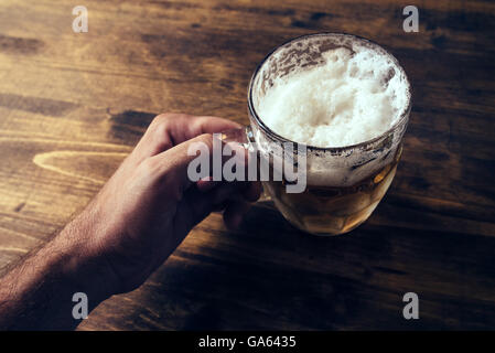 Hand holding beer mug full of cold fresh alcohol drink on wooden background, man in the bar, pov shot, selective focus Stock Photo