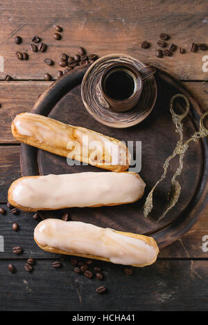 Fresh homemade eclairs with different glaze, coffee beans and ceramic cup of black coffee over old wooden texture background with space for text. Top Stock Photo