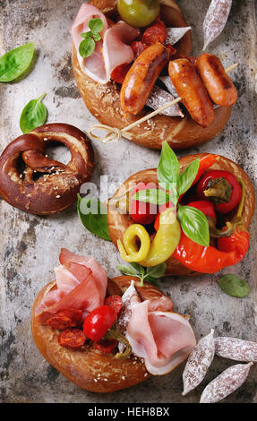 Variety of meat snacks fried sausages, wienerwurst, ham, marinated chili peppers served in salted pretzels with fresh basil over old metal background. Stock Photo