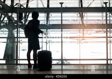 Silhouette of a traveler with luggage suitcase and backpack look out of the window at airport Stock Photo