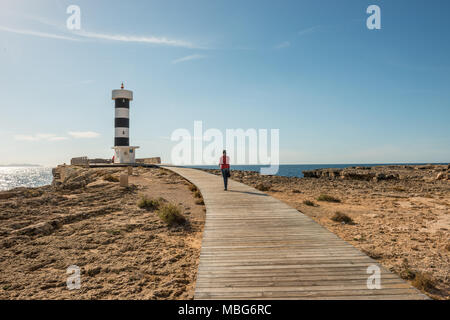 A young woman with gray hair and a red scarf walks along the walkway towards the lighthouse on the coast in Colonia de Sant Jordi, Mallorca Island. Stock Photo