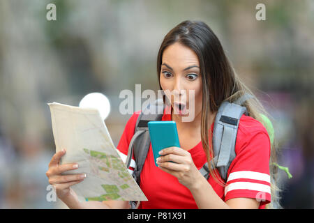 Surprised teen tourist holding a map and reading online content in a smart phone on the street Stock Photo