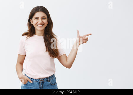 Portrait of enthusiastic helpful and attractive young european woman with brown long hair holding hand in pocket of jeans and pointing right with finger gun gesture smiling broadly showing way out Stock Photo