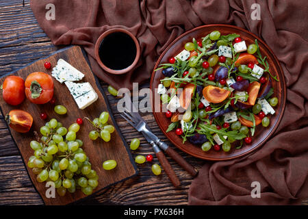 delicious autumn salad with chocolate persimmon, blue cheese mold, green grapes, plum slices and arugula on a earthenware plate with ingredients on a  Stock Photo