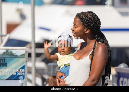 Cape Town, South Africa - January 2th, 2019: A south african black young mother with her baby saying hello in her arms at the Cape Town waterfront. Stock Photo